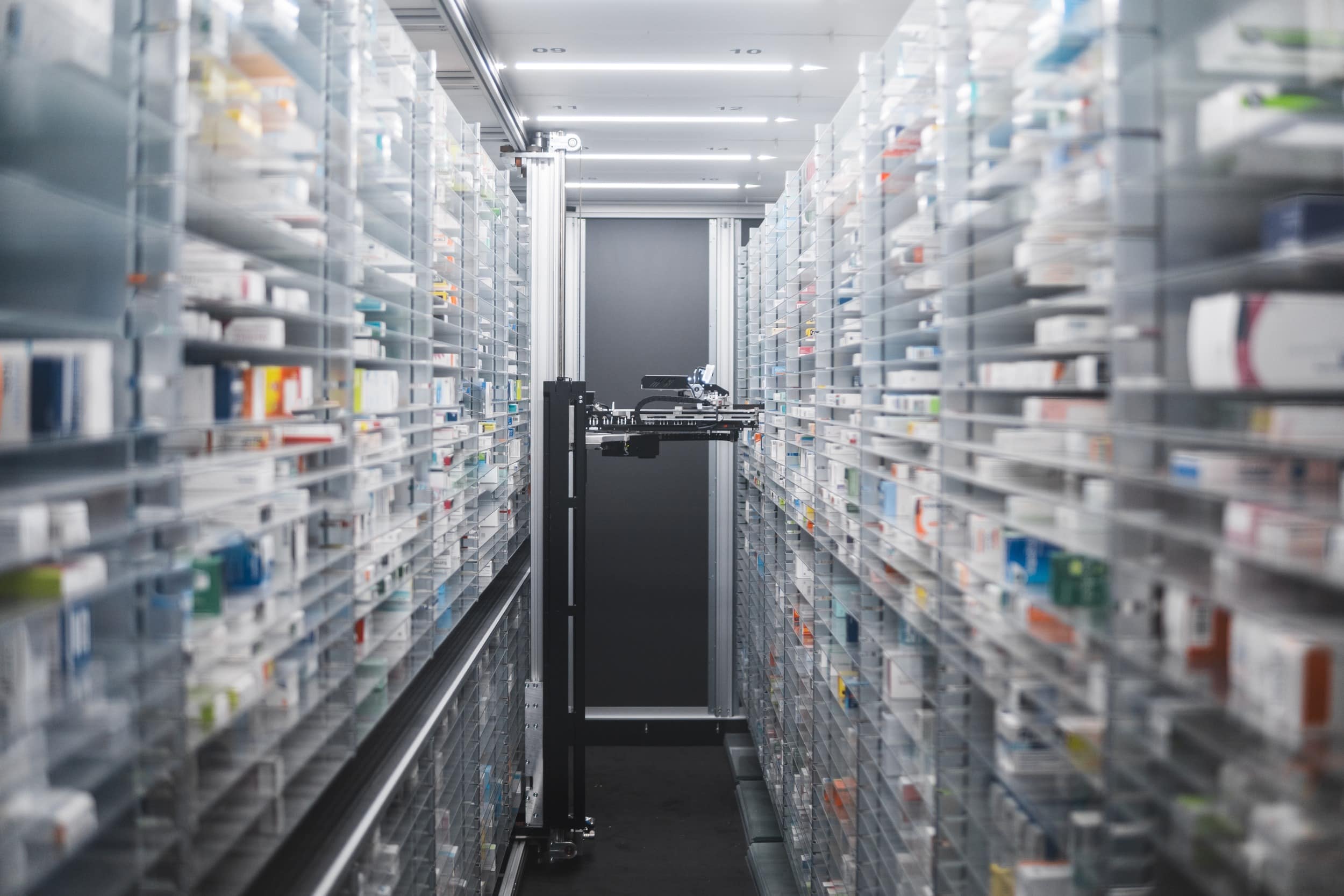 Narrow pharmacy aisle flanked by towering shelves stocked with medicine boxes. A pharmacy robot navigates through, ensuring efficient and precise medication retrieval.