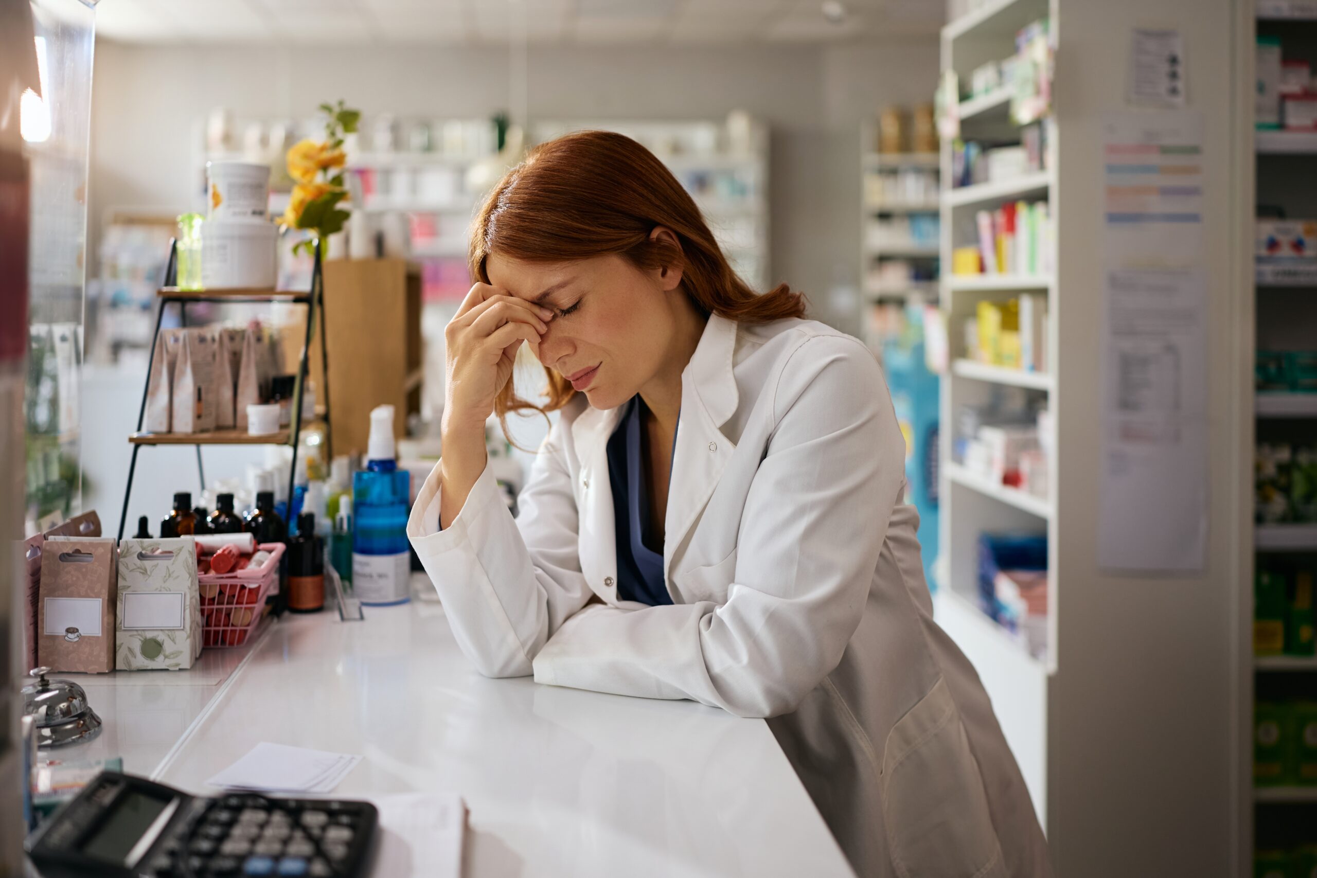 A pharmacist rests her head in her hand at the pharmacy counter, looking stressed.
