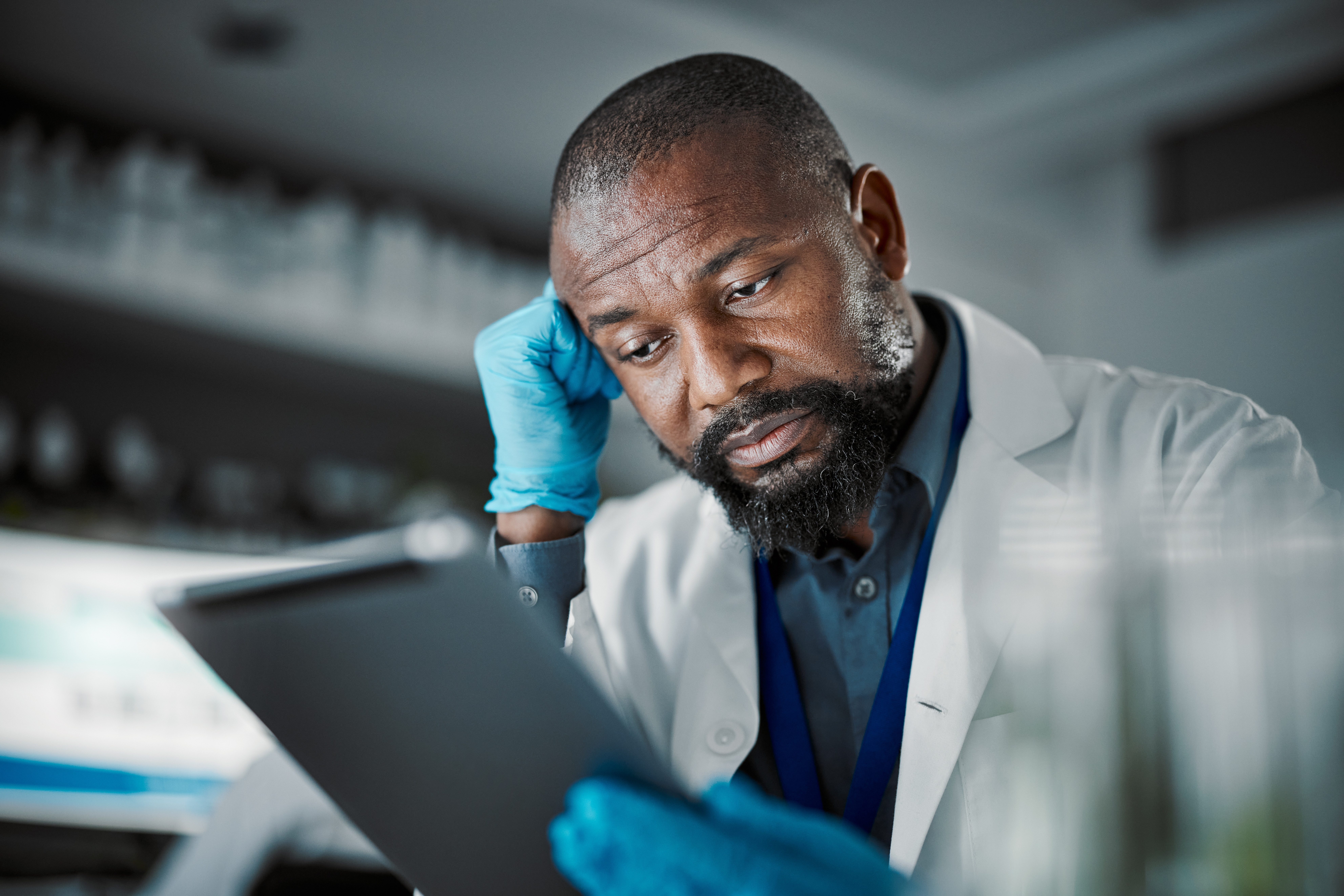 A lab worker in gloves and a white coat looks tired while reviewing information on a tablet.