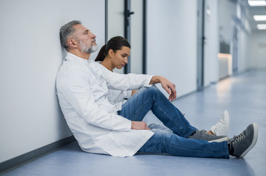 Two pharmacy workers sit on the floor in a hallway, appearing tired or overwhelmed.