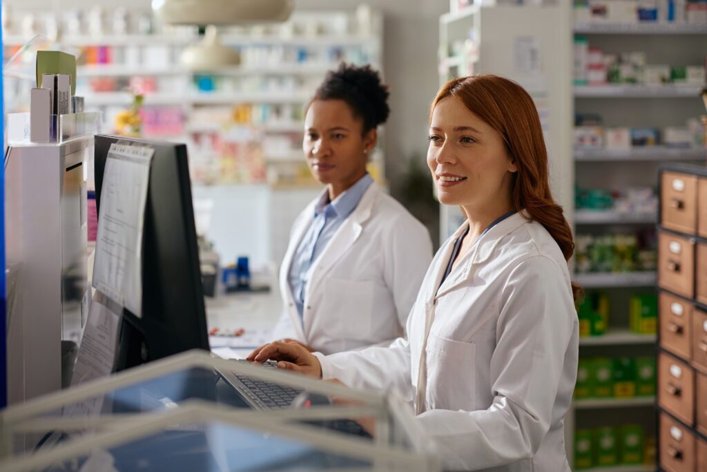 Two pharmacists in white coats work at a computer behind the pharmacy counter.