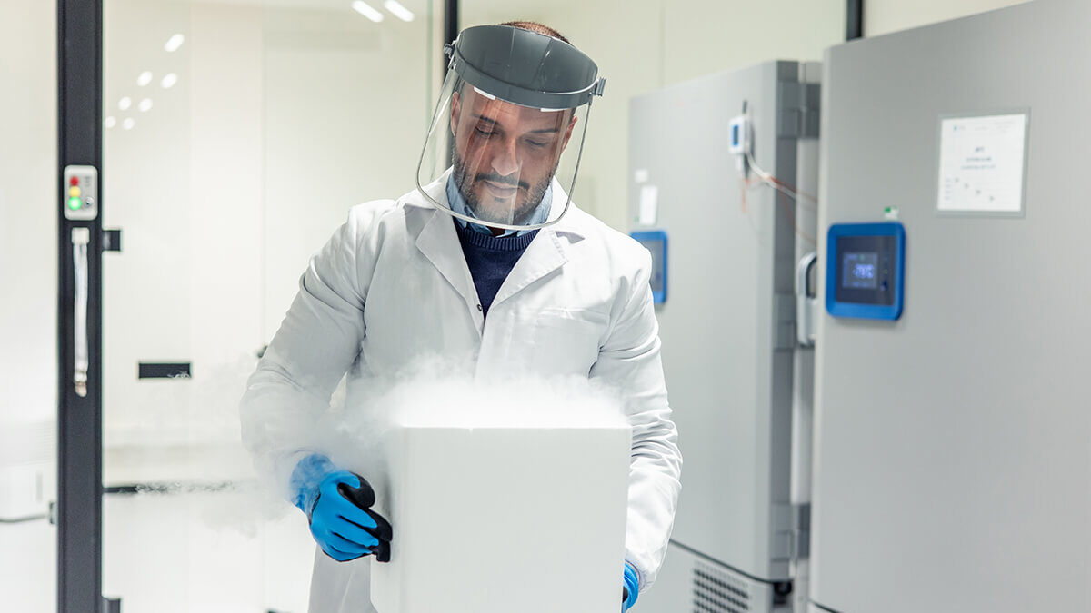 Researcher in a lab coat and face shield handling a steaming cryogenic storage container in a medical laboratory.